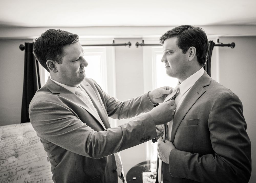 Black and white image showing two people in suits adjusting each other's neckties in a formal setting.