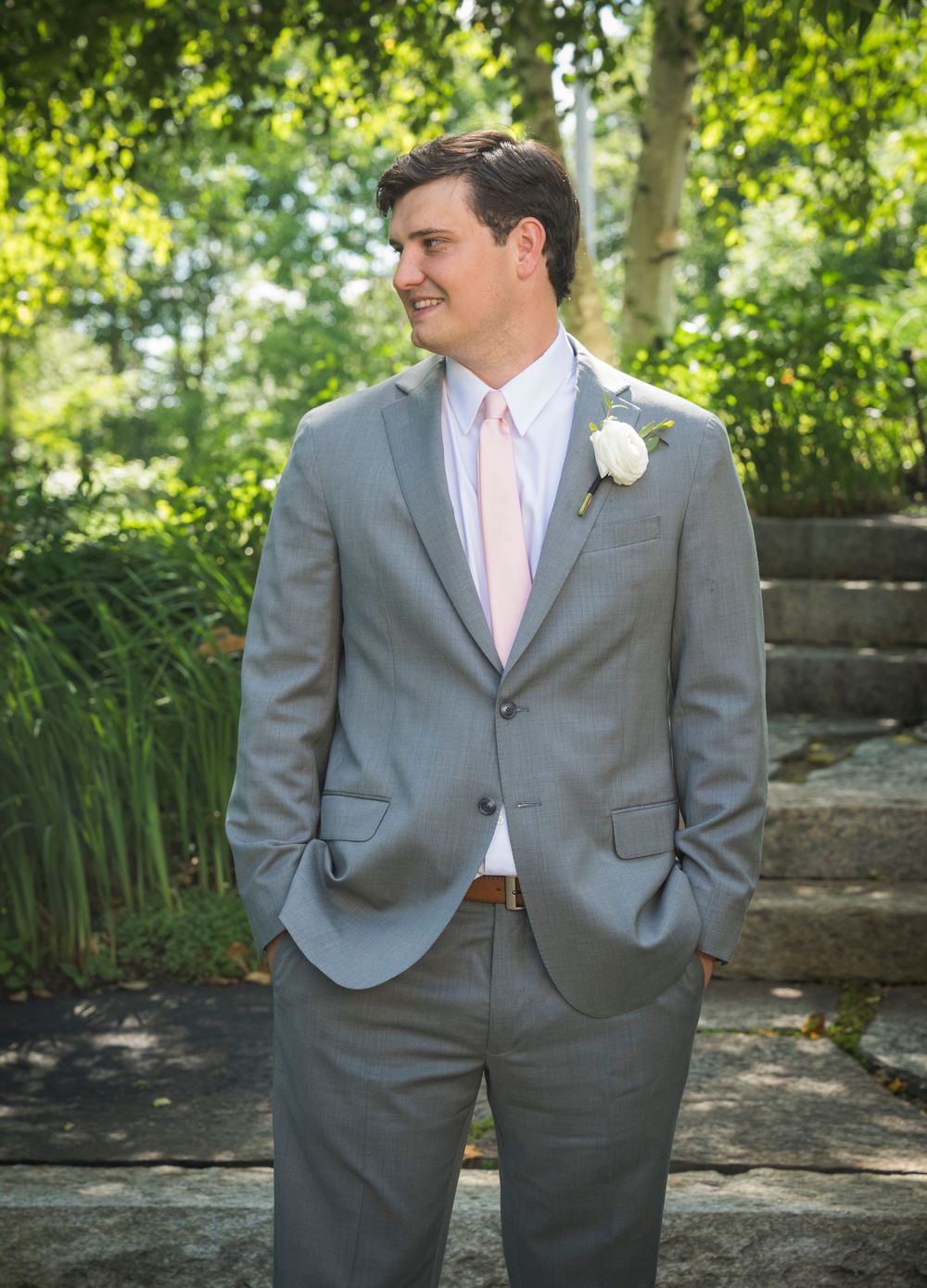 A groom in a light grey suit and pink tie poses outdoors in a garden setting.