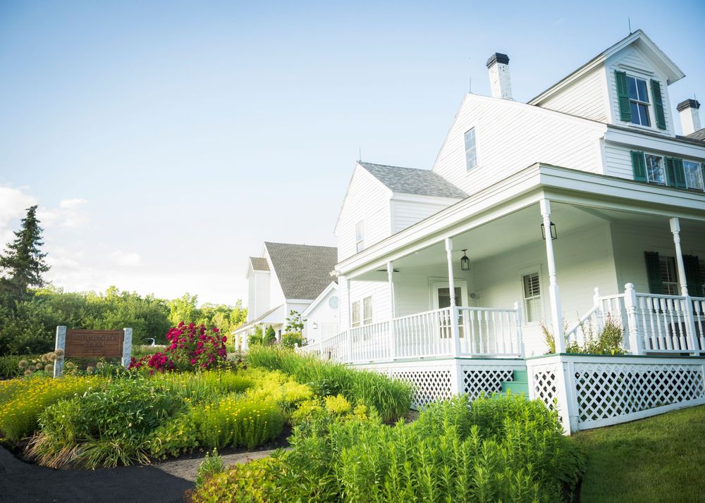 White farmhouse with wraparound porch and colorful garden in full bloom against blue summer sky.