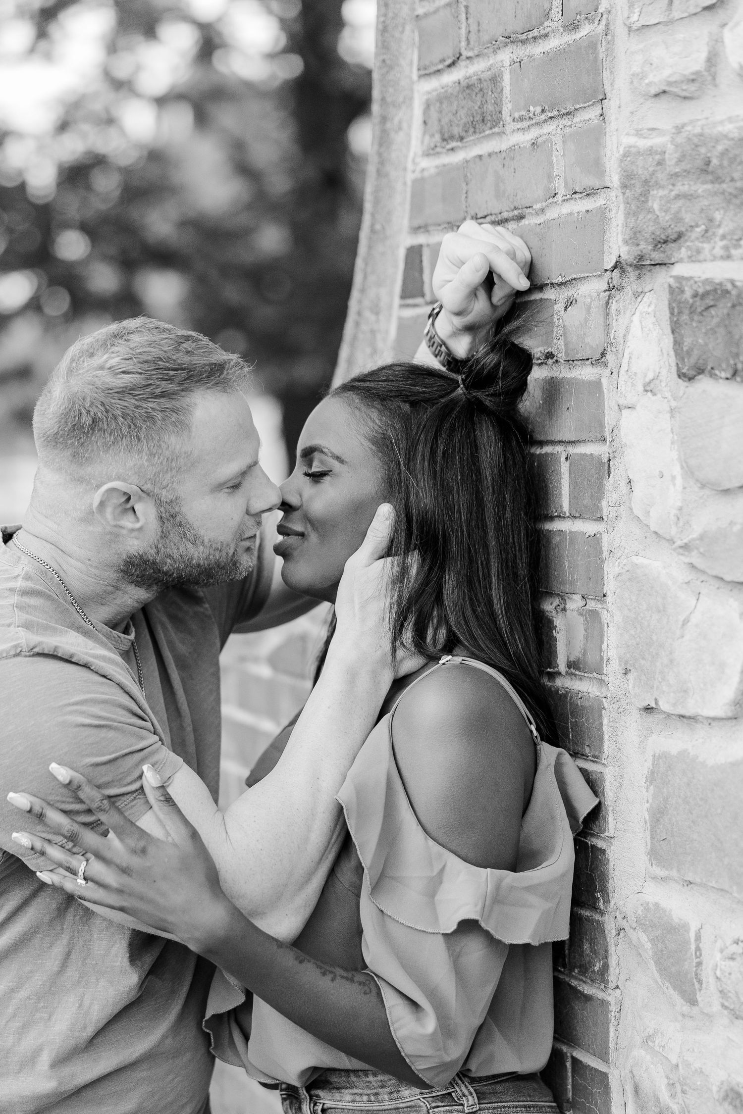 A romantic couple shares a tender embrace against a brick wall in this black and white portrait.