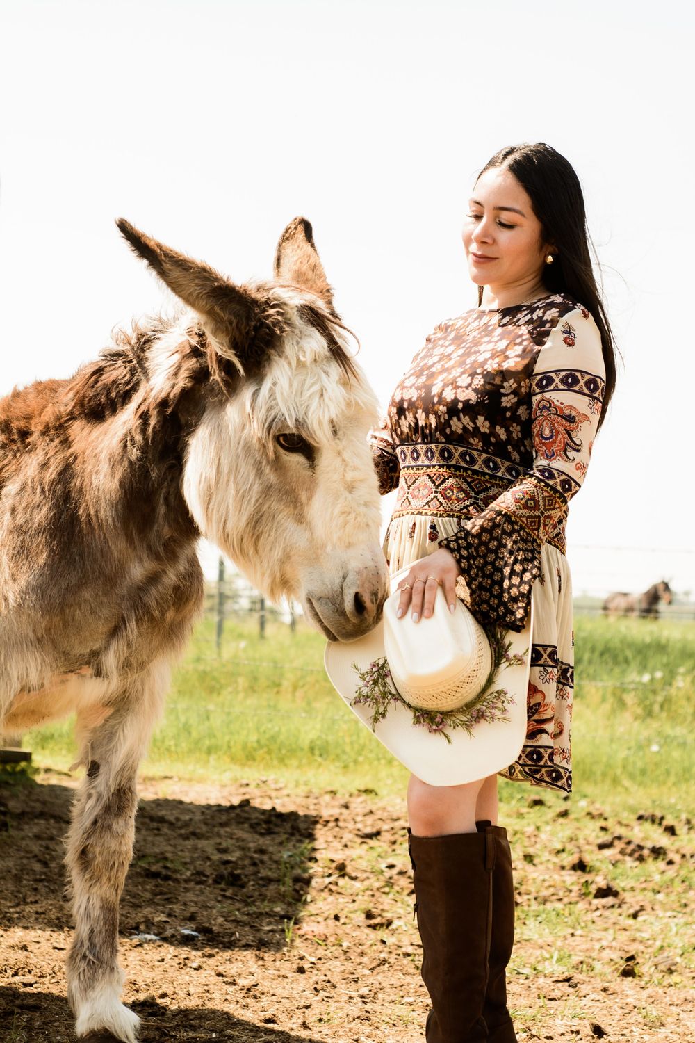 A person in a patterned dress stands next to a friendly donkey in a rural field setting.