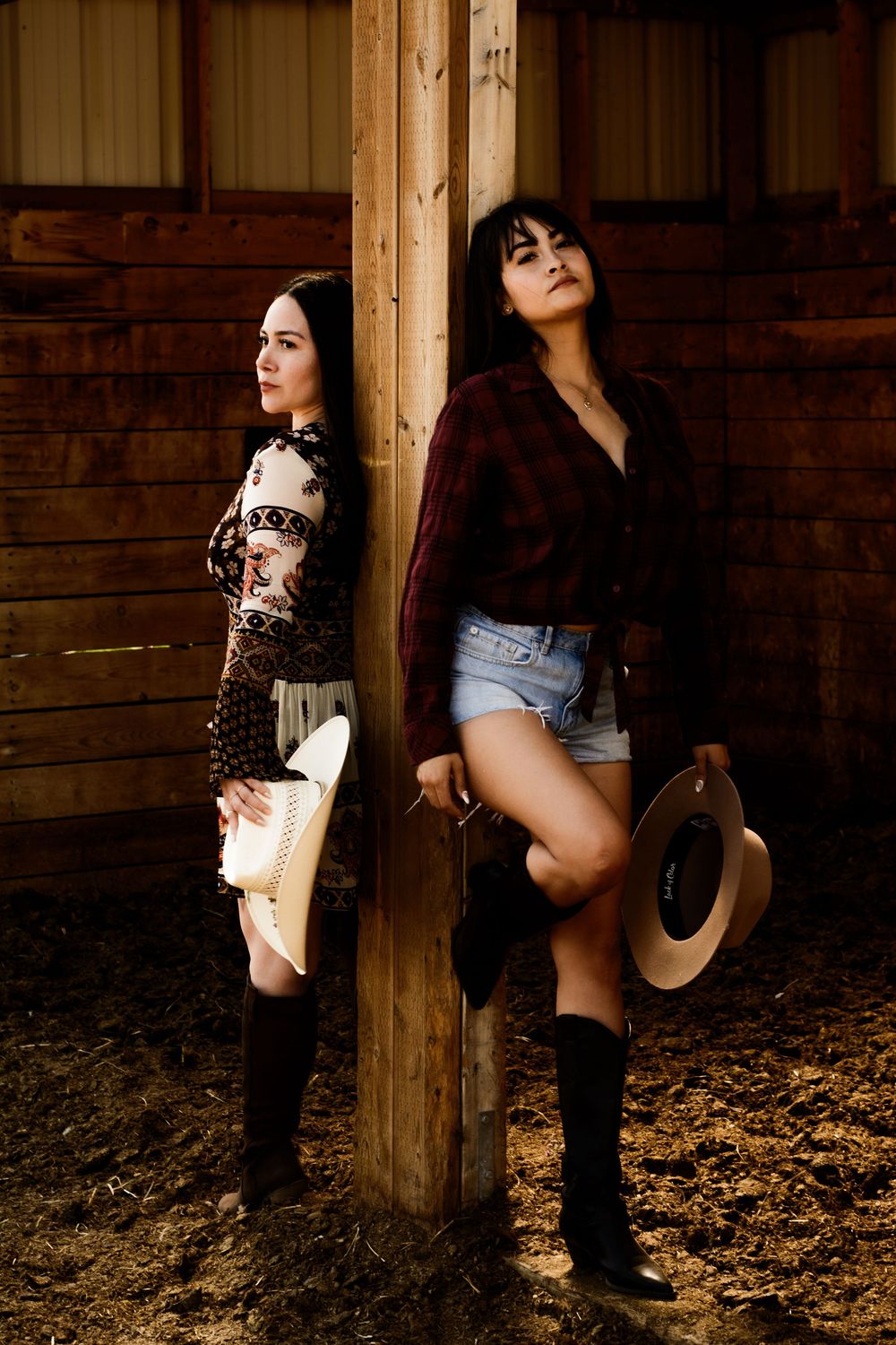 Two women in western outfits pose against wooden barn posts.