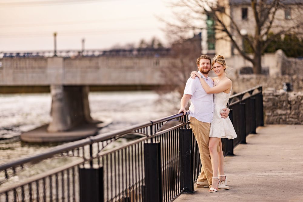 Couple standing together on riverwalk promenade with bridge and railing during sunset.
