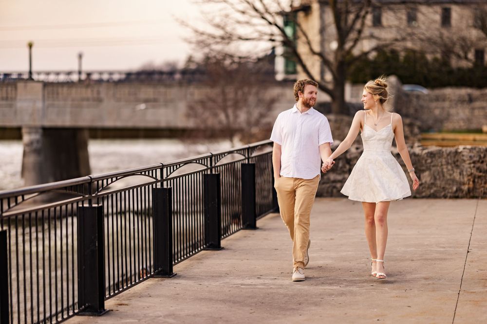 A couple walking together along a waterfront path with a bridge in the background on a sunny day.