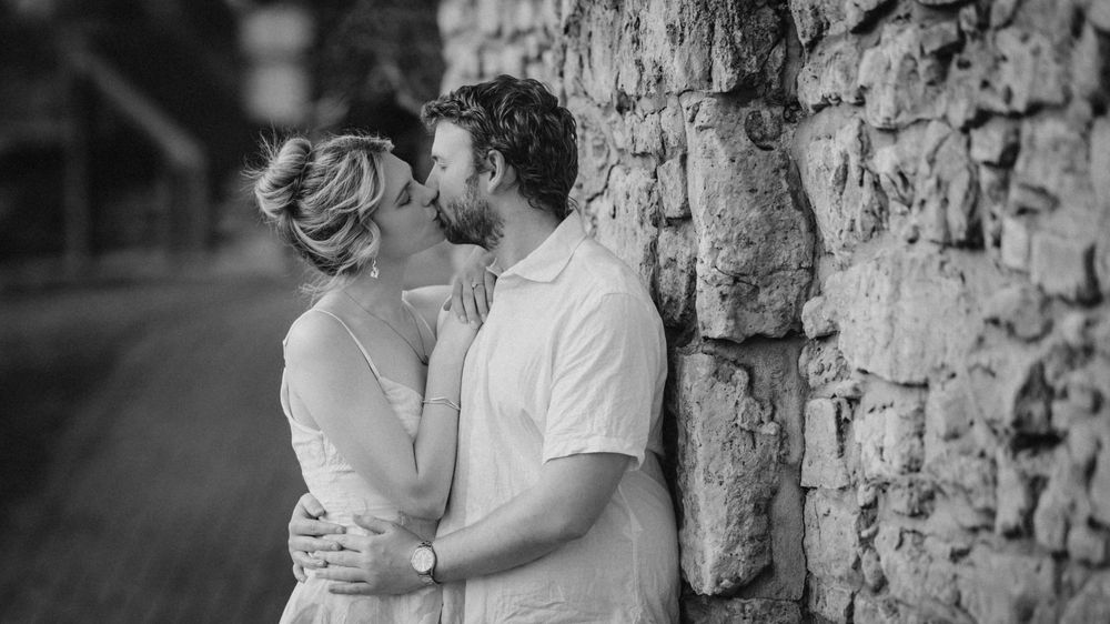 A romantic black and white image showing a couple in a tender embrace against a rustic stone wall.