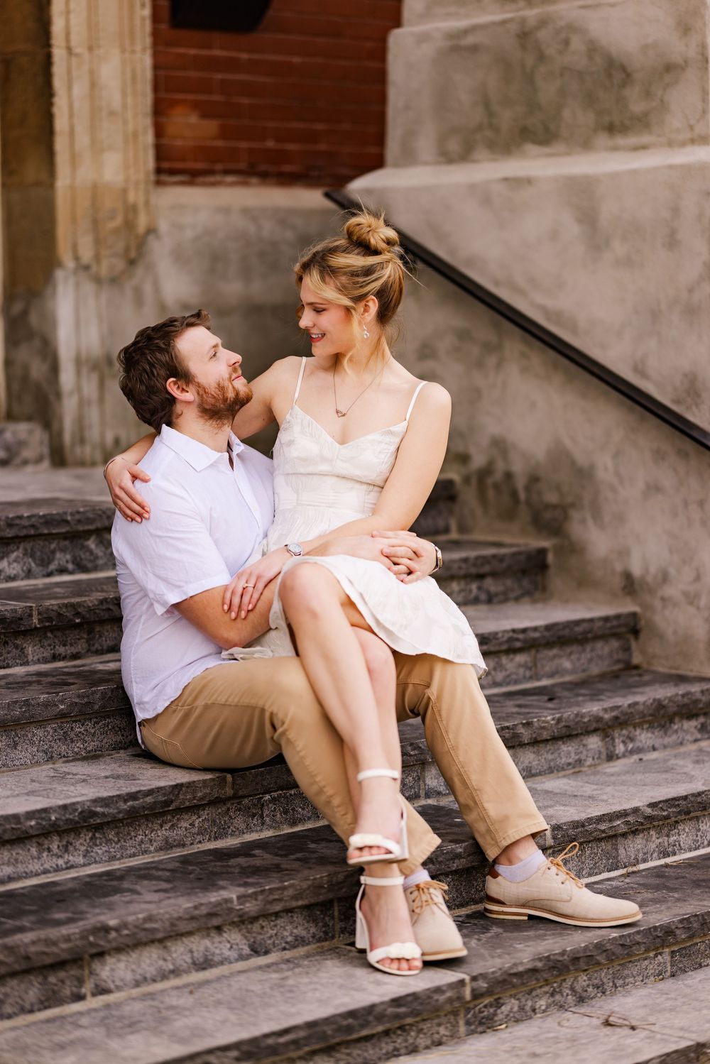 A couple sits intimately on stone steps, wearing casual summer attire with a vintage architectural backdrop.