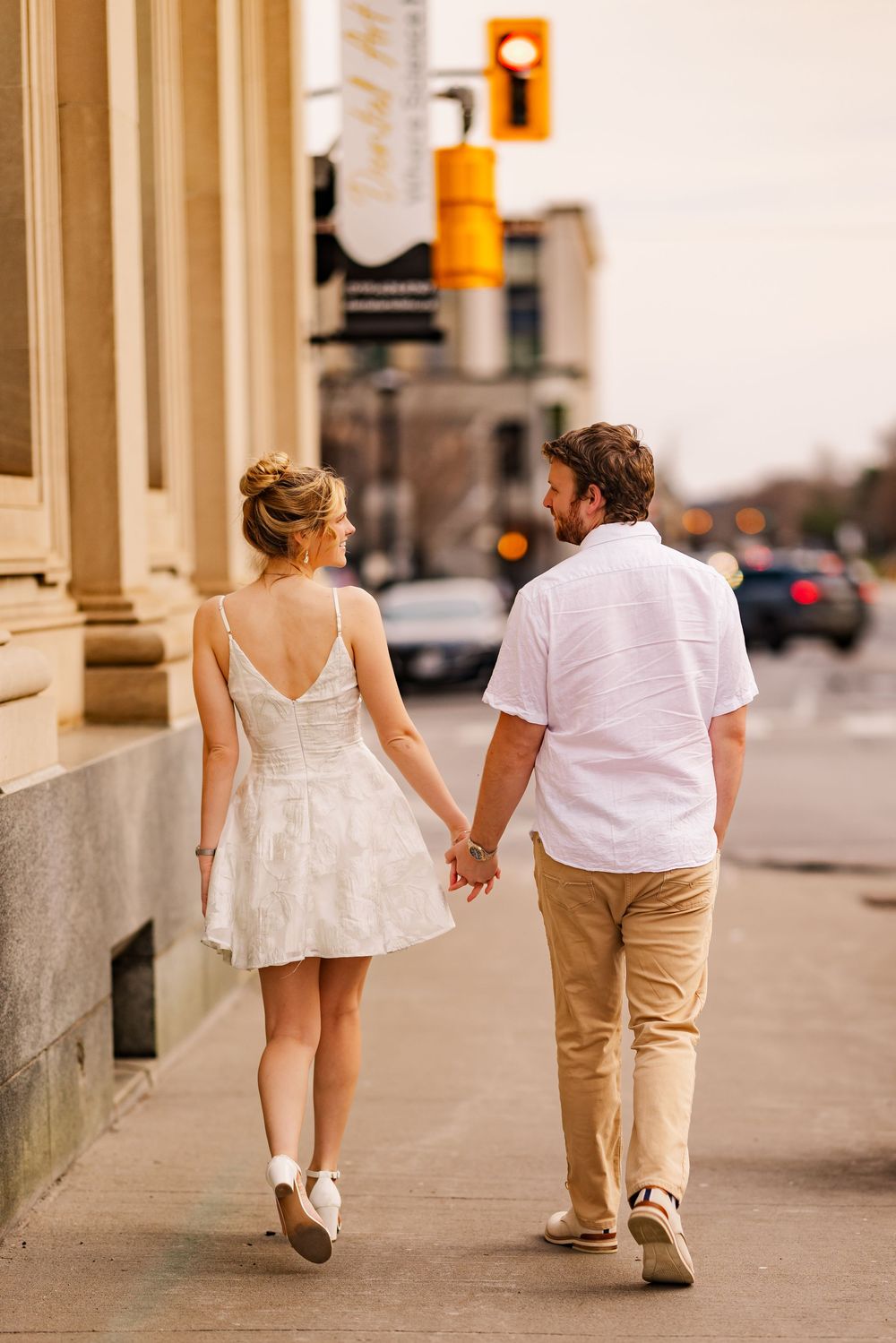 Young couple holding hands while walking on a city sidewalk, with a traffic light visible in the background.