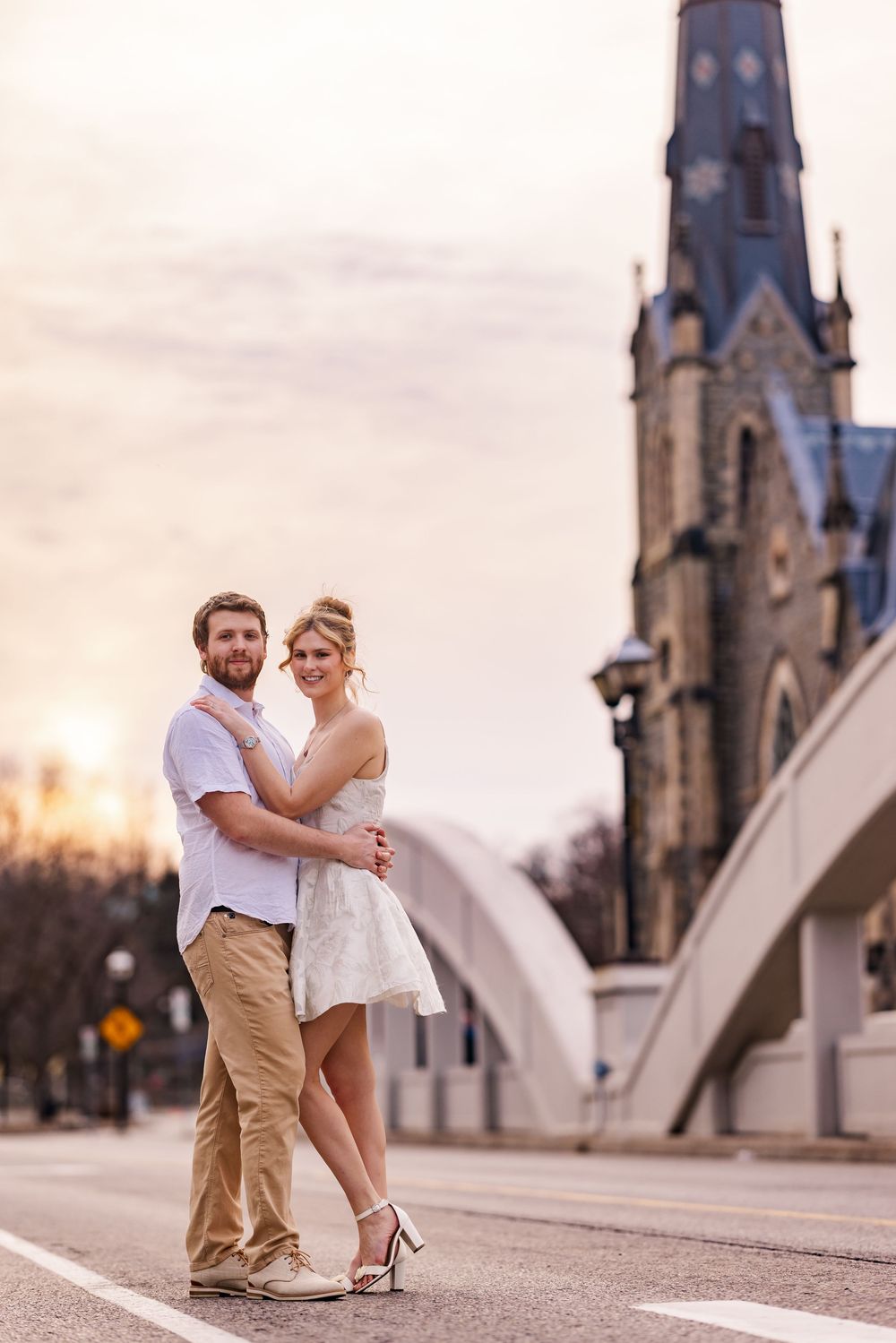 A couple embraces in front of a Gothic church spire during sunset on a city street.