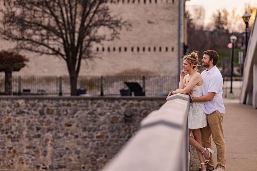 A couple stands close together at sunset along a stone wall and railing with historic architecture in the background.