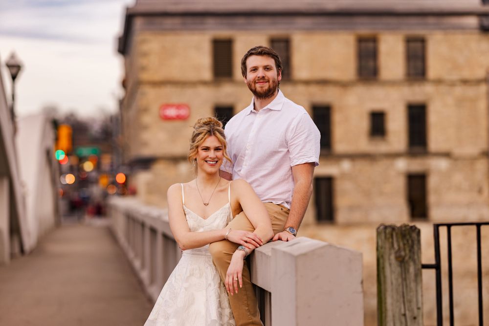 A couple in wedding attire pose together on a concrete barrier near an old building during sunset.