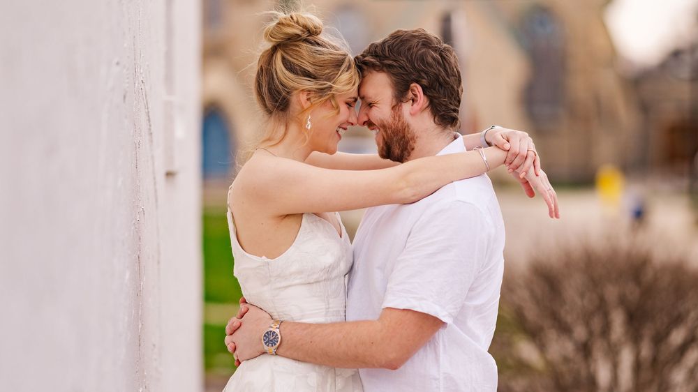 A loving couple in casual attire embrace intimately outdoors against a blurred architectural backdrop.