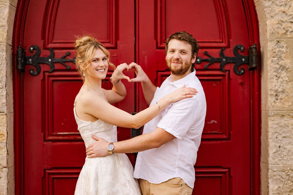 A couple poses making a heart shape with their hands in front of a vibrant red wooden door.