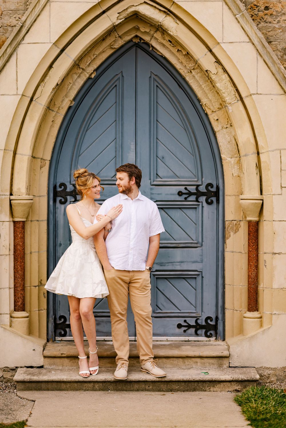 A couple in white outfits poses playfully in front of medieval stone archways with blue doors.