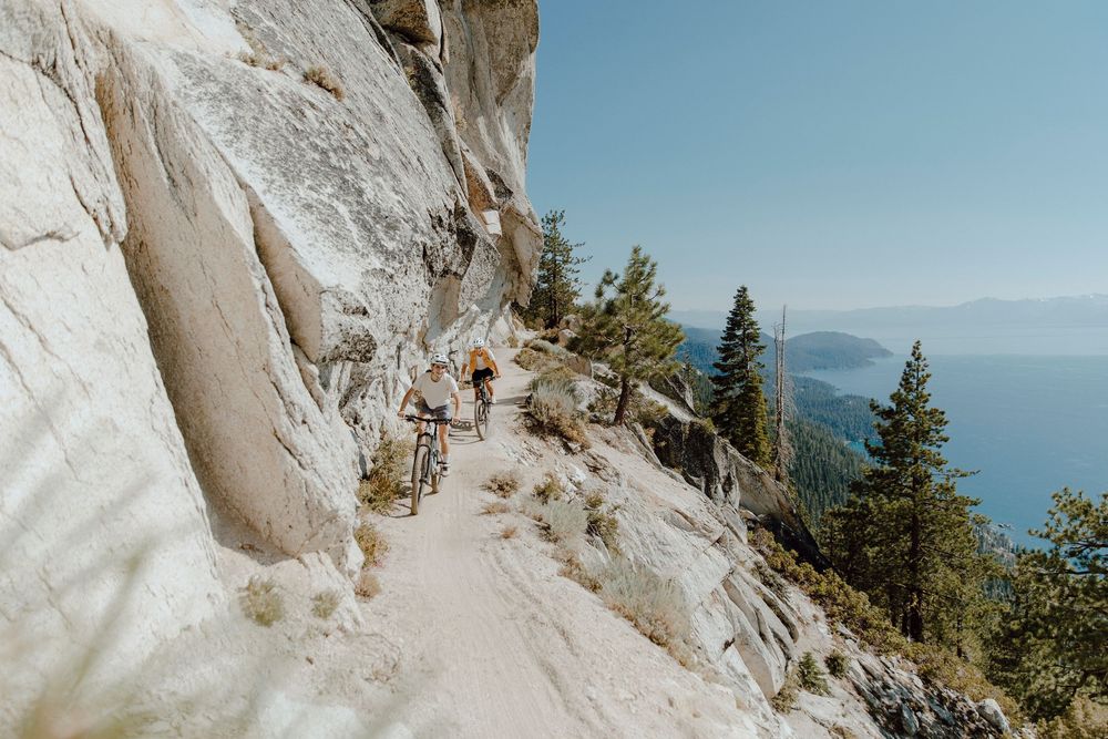 Hiking trail along steep granite cliffs overlooking Lake Tahoe with evergreen trees and blue sky in the background.