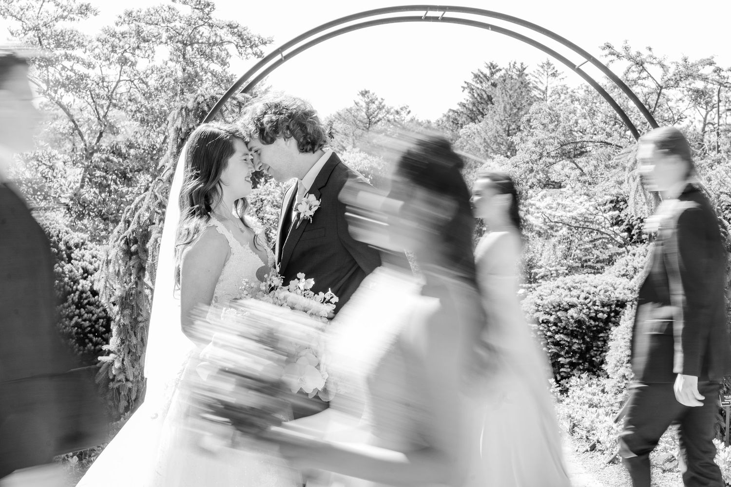 A wedding couple stands under an arch while guests walk past in a motion blur effect at an outdoor garden ceremony.