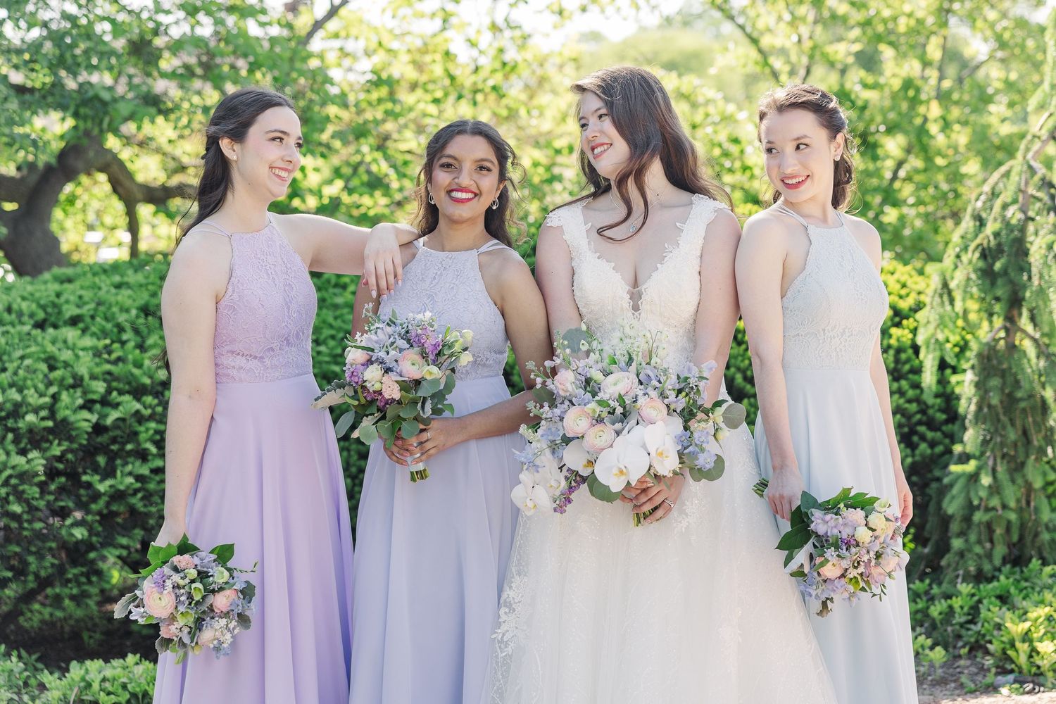 A bride and bridesmaids in pastel purple dresses hold floral bouquets while standing together outdoors in a garden setting.