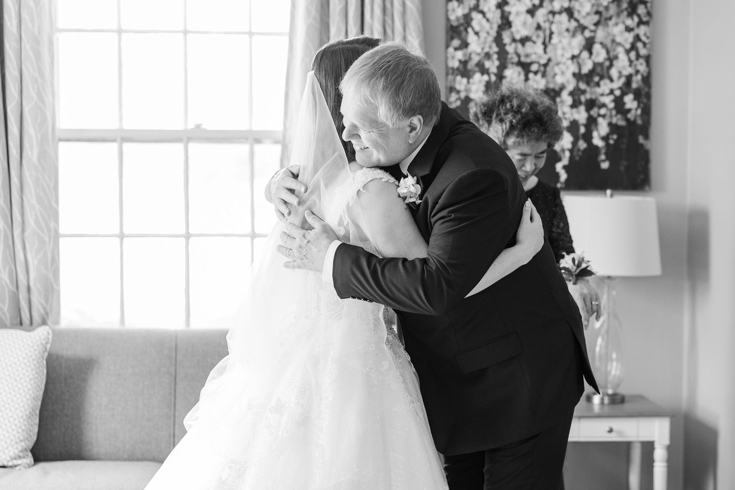A black and white wedding photograph captures an emotional embrace between people in formal attire near a window.