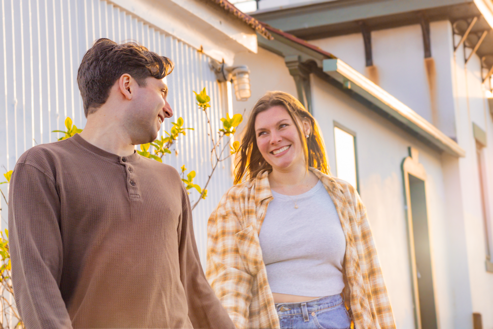 A couple enjoying a sunny day together in front of a house with yellow flowers in the background.