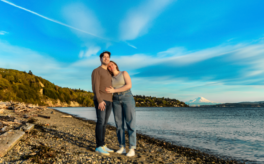 A couple poses together on a rocky beach shoreline with mountains and blue skies in the background.