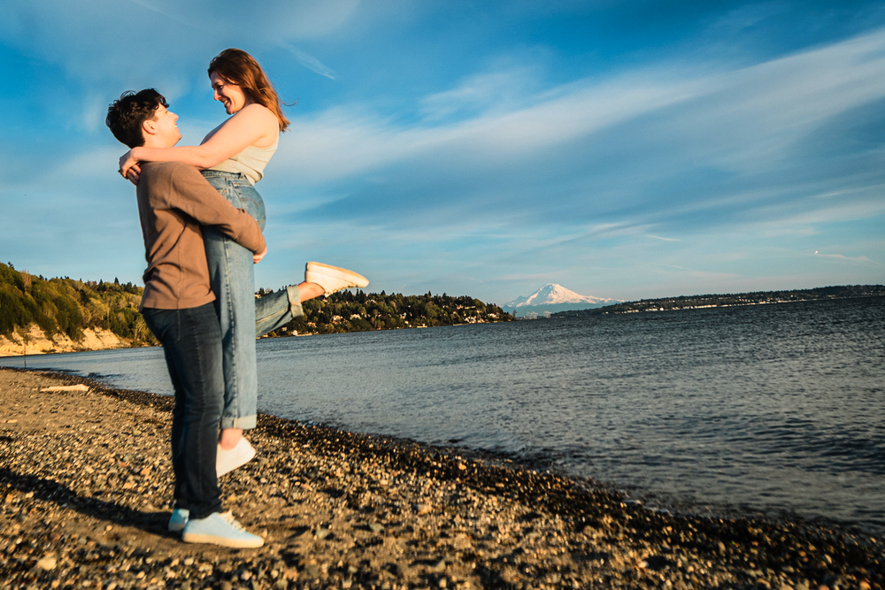 A romantic couple embraces on a rocky beach during golden hour with calm waters and a blue sky in the background.