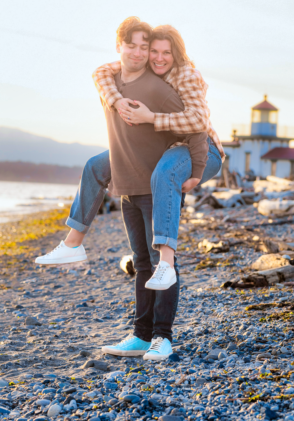A couple shares a playful moment on a rocky beach at sunset with a lighthouse in the background.