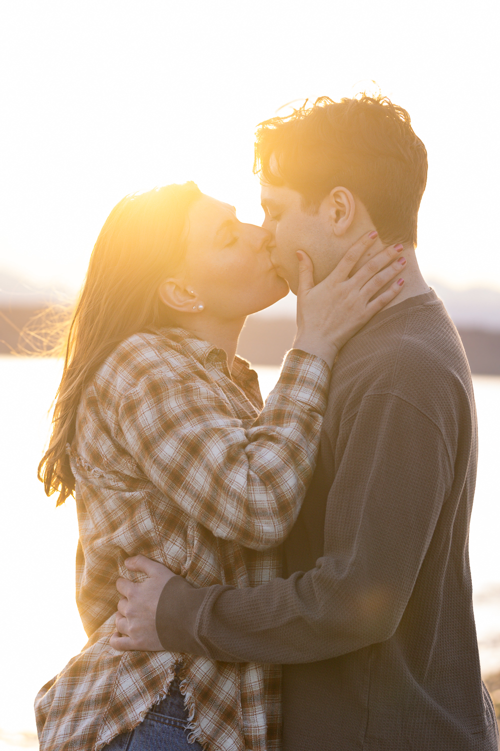 A romantic couple shares a tender kiss at sunset with golden light streaming behind them wearing plaid and dark clothing.