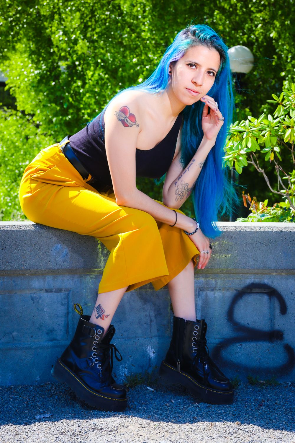 Alternative fashion portrait with bright blue hair, black top, yellow pants and chunky platform boots against green foliage.