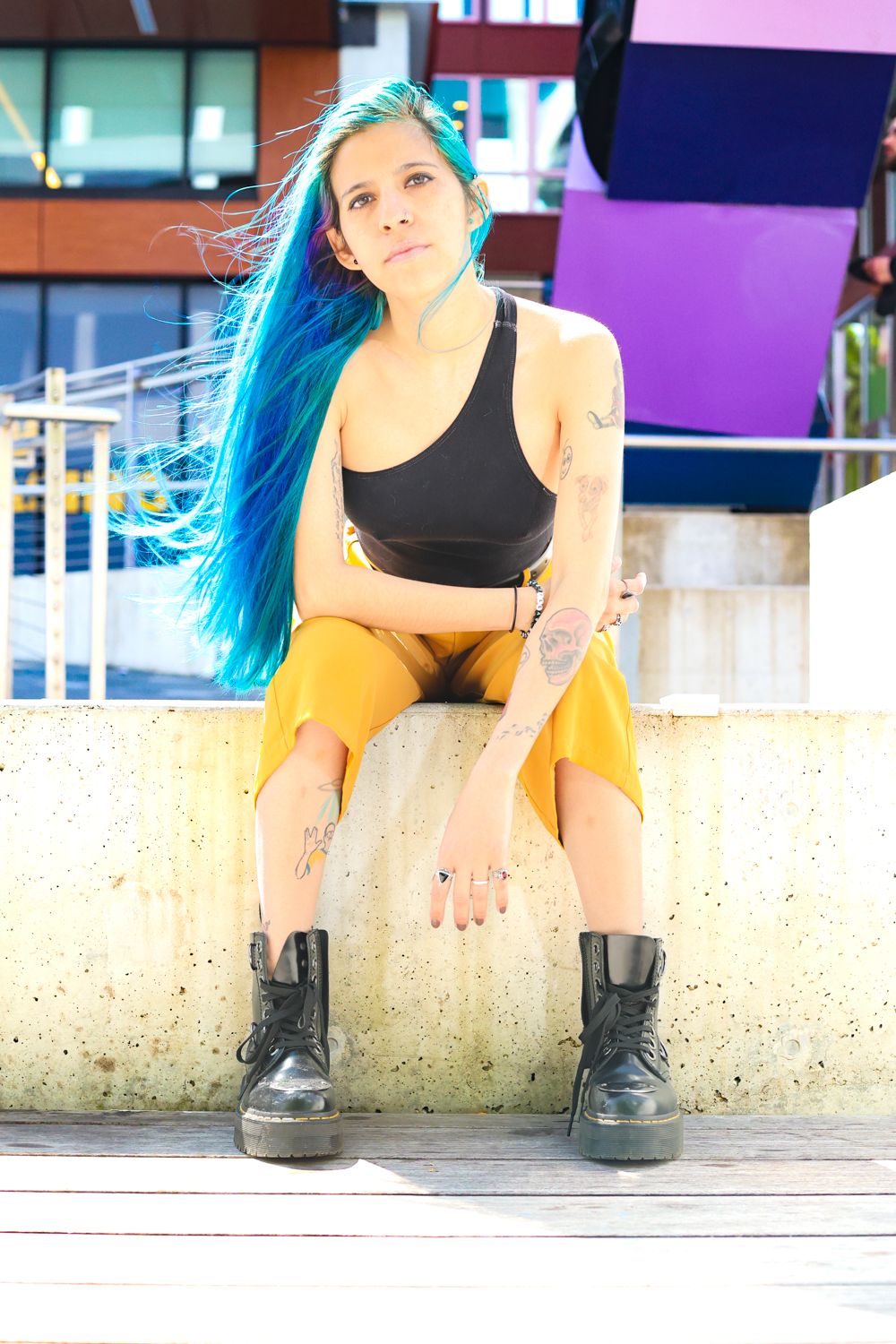 A person with vibrant blue hair wearing black top and yellow pants sits on a concrete ledge.