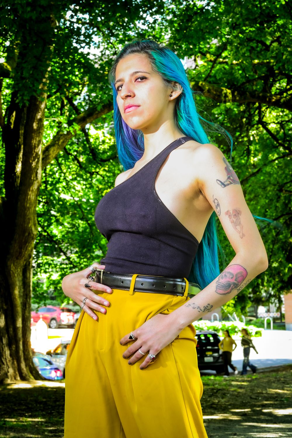 A person in a black tank top and yellow culottes poses against a lush green park background with visible tattoos.