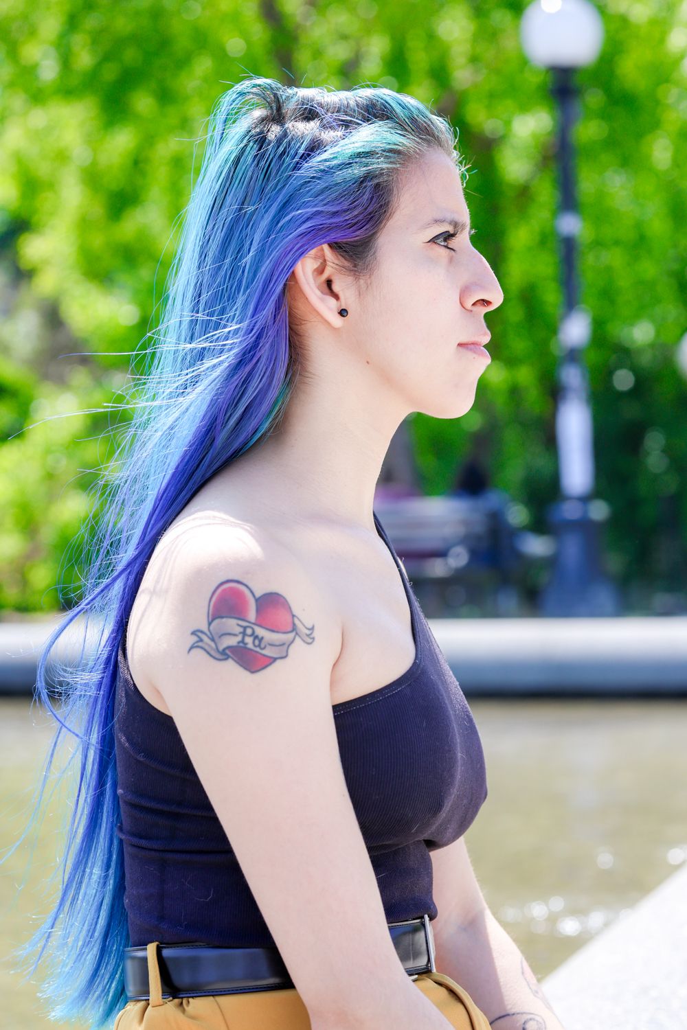 Side profile of a person with vibrant blue and purple ombre hair wearing a black top outdoors near a lamp post.