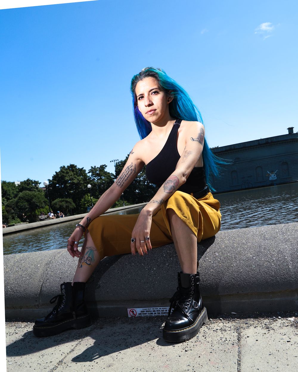 Person with bright blue hair wearing a black top and yellow pants sitting casually on a stone ledge near water.