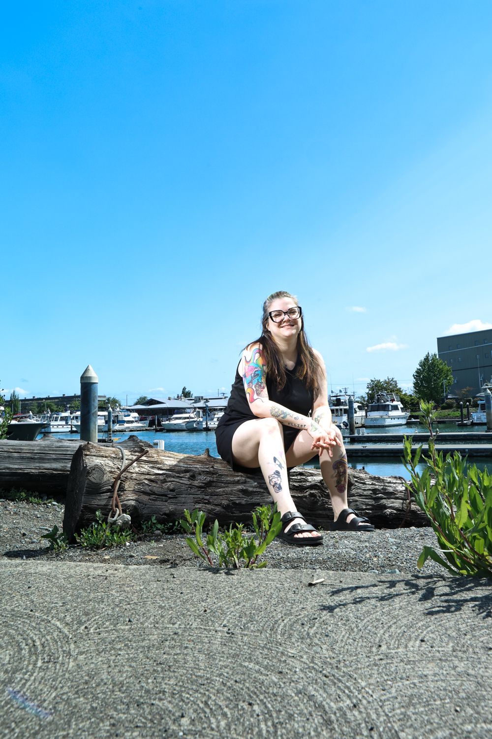 A person in casual wear sits on a log at a marina with boats and blue sky in the background.