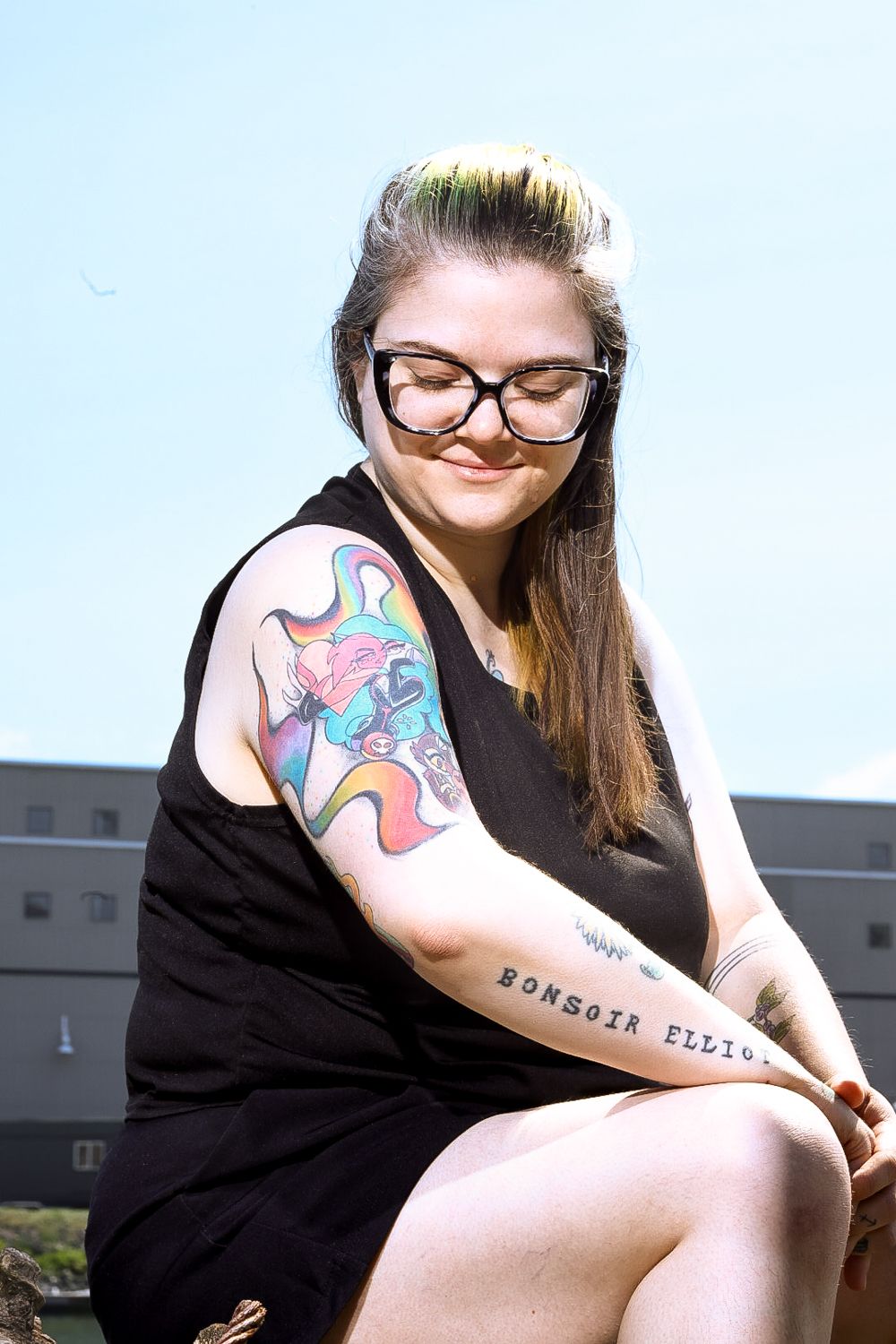 A person in glasses and a black dress shows off a colorful abstract tattoo on their upper arm.