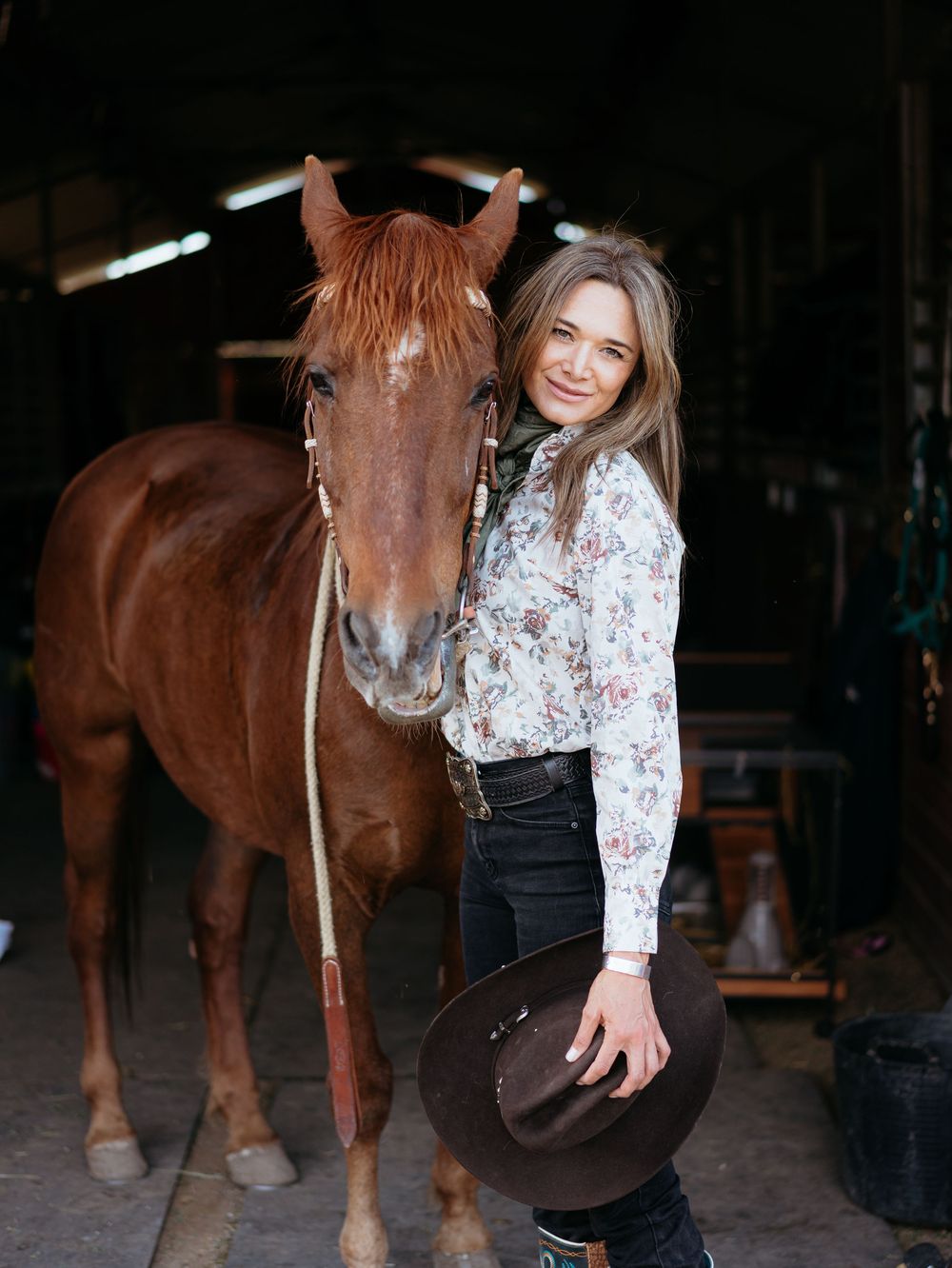 A person stands with a chestnut brown horse in a rustic barn while wearing a floral blouse and dark jeans.