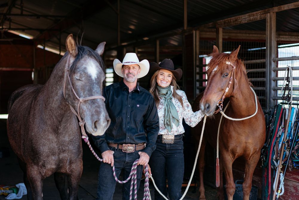 Two people in western wear stand between two horses inside a barn with equestrian equipment visible.