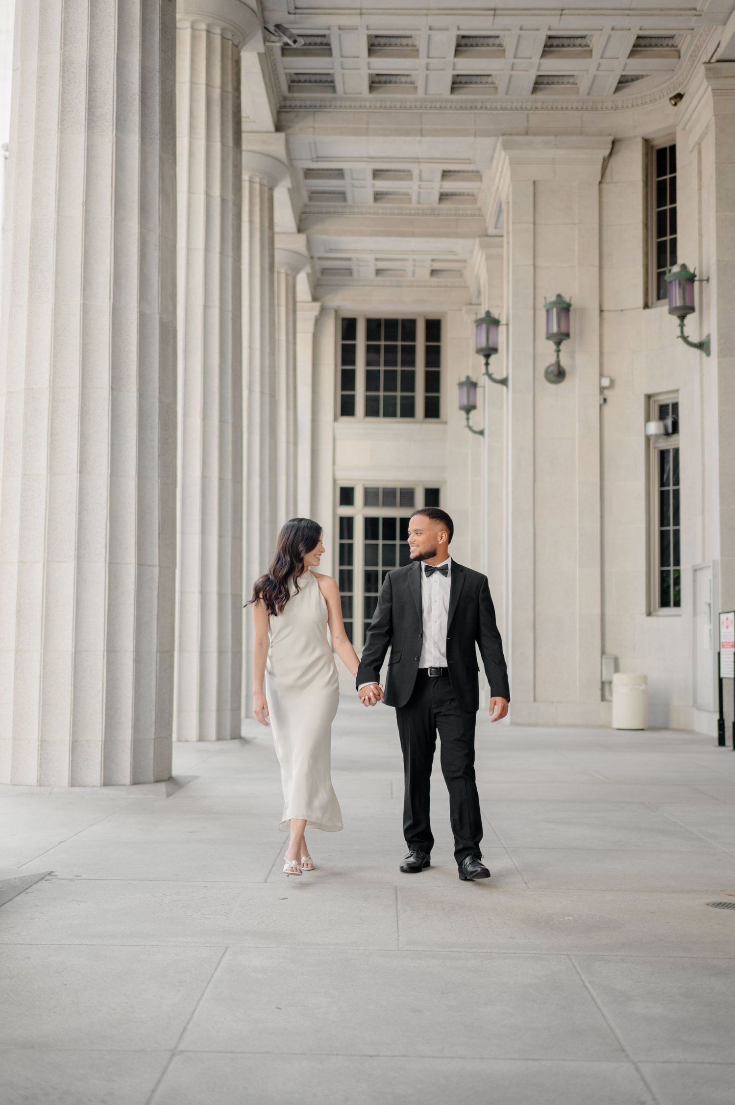 A couple in formal attire walks hand in hand through an elegant colonnade with classical white columns and sconces.