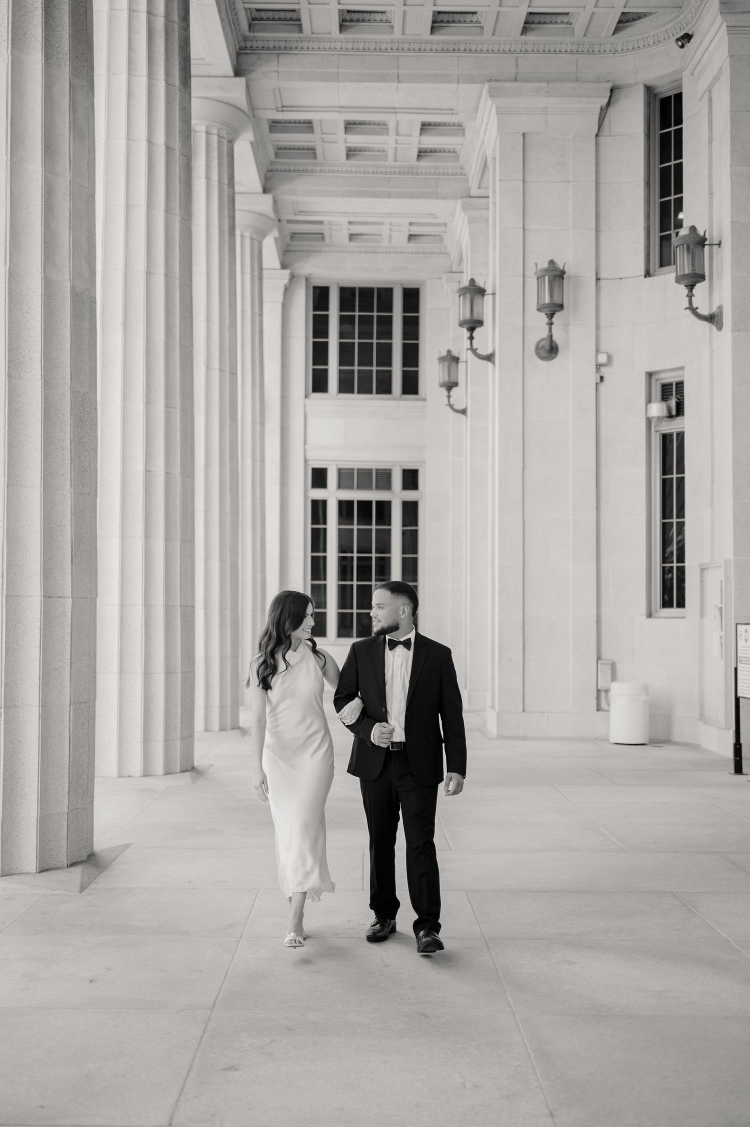 A couple in formal attire walks together beneath elegant white columns and lanterns at a grand venue.