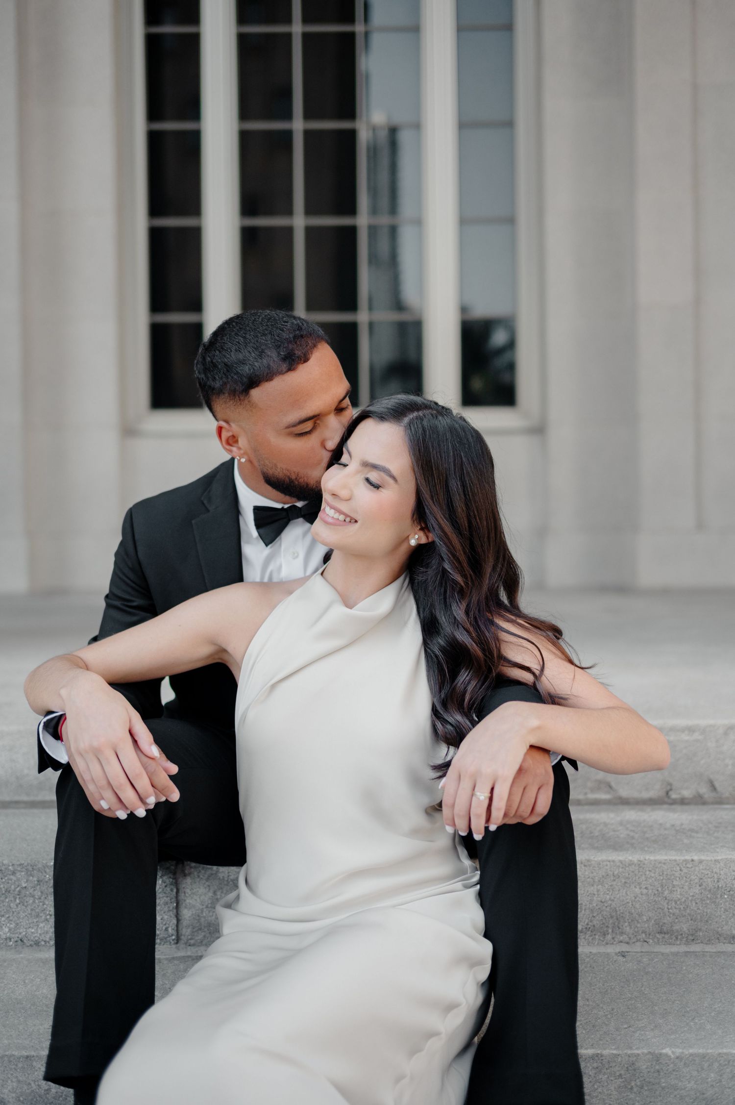 Couple in formal wear embraces on steps against architectural backdrop.