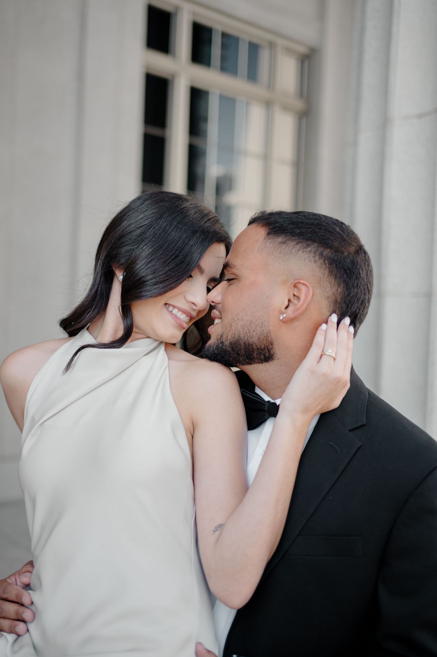 Romantic couple in formal attire share an intimate moment against a white wall with a window frame backdrop.