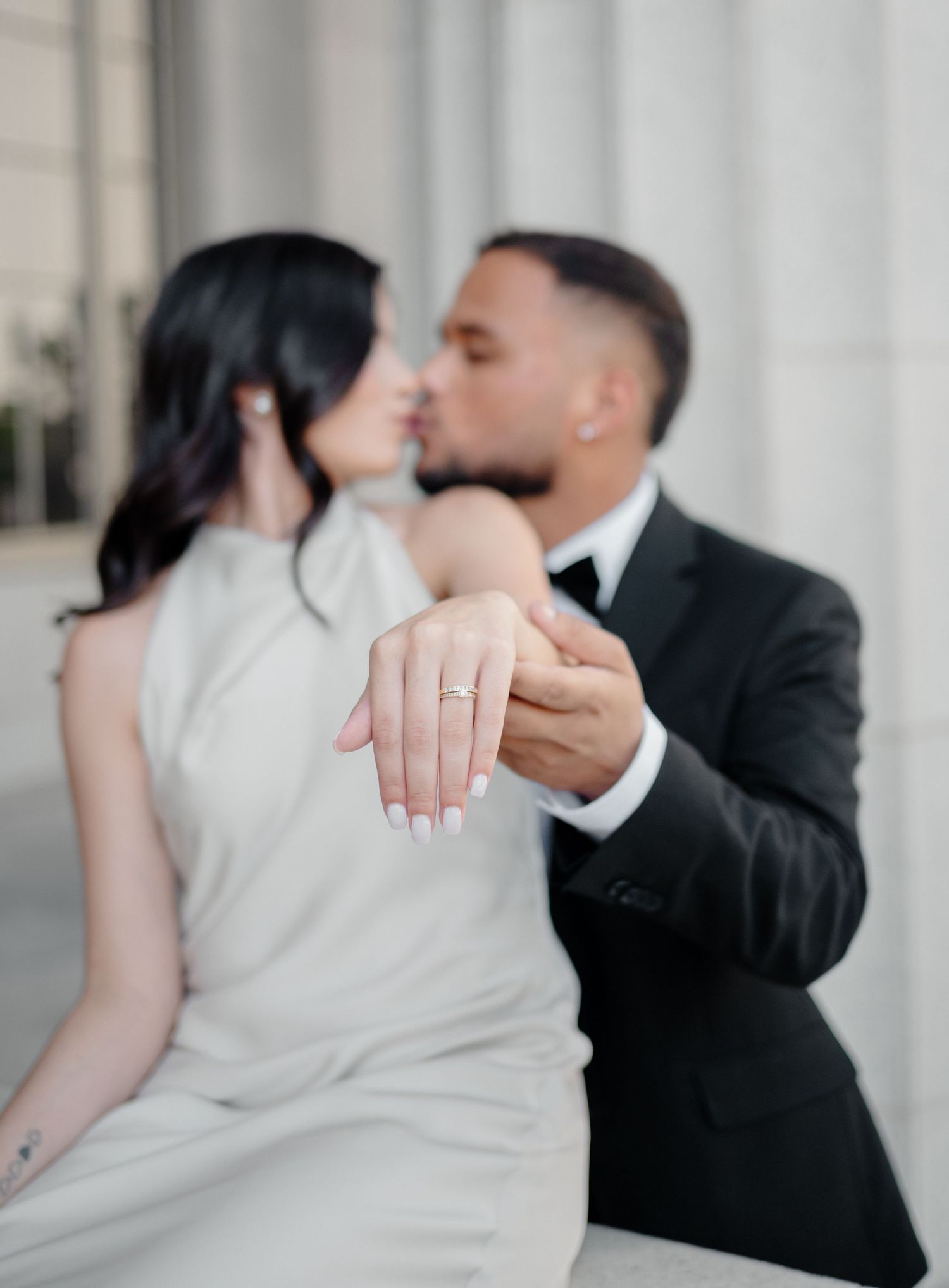 A romantic couple shares a tender moment in front of white columns while displaying an engagement ring.