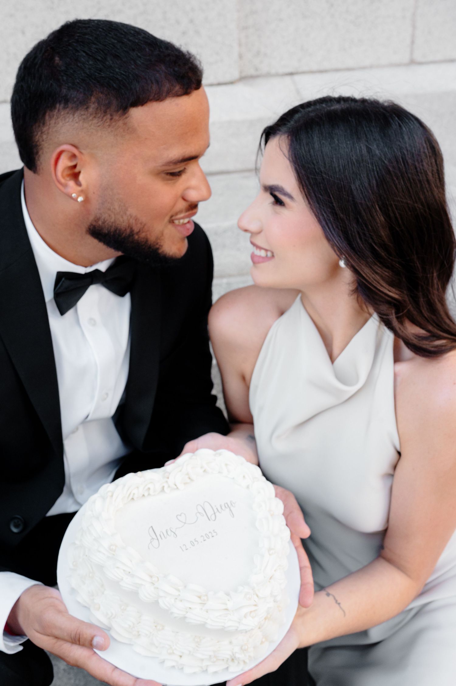 Elegant couple holds white heart-shaped wedding cake while gazing lovingly at each other in formal attire.