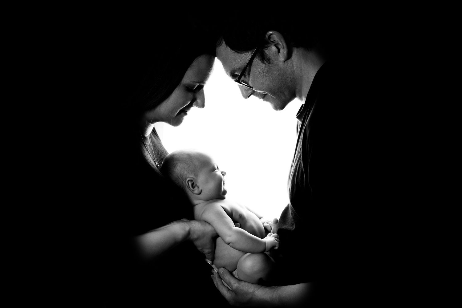 A tender black and white portrait showing an emotional moment between parent and baby lit dramatically against dark shadows.