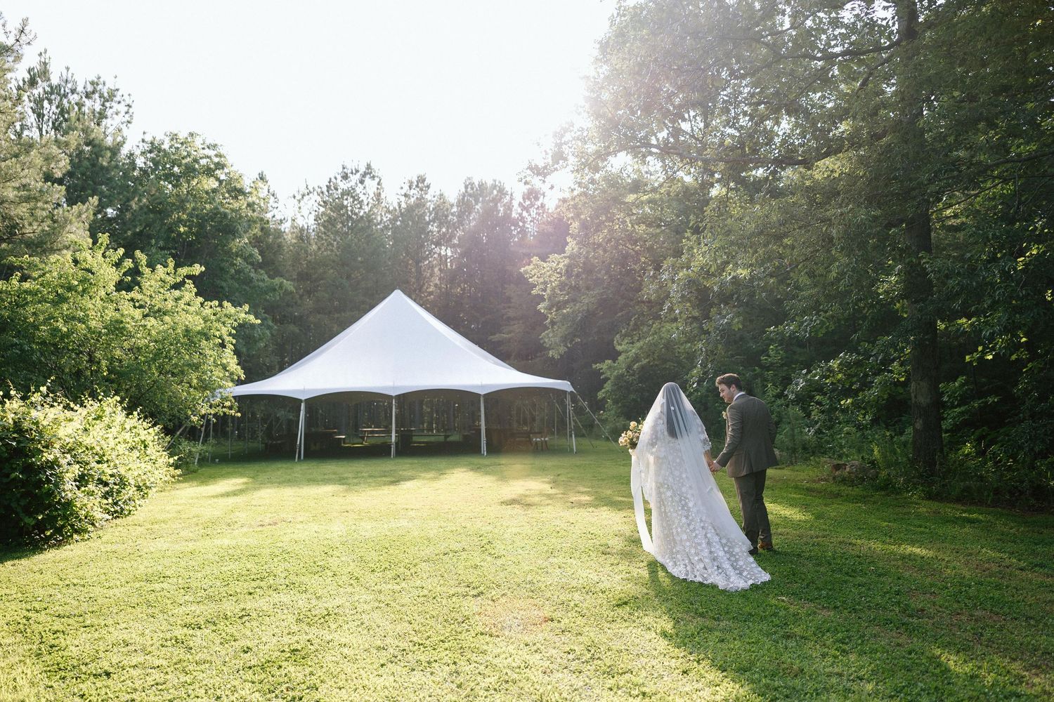 Couple walking on grassy lawn towards white wedding tent during sunset in outdoor garden setting at The House Boheme in Lake Lure.