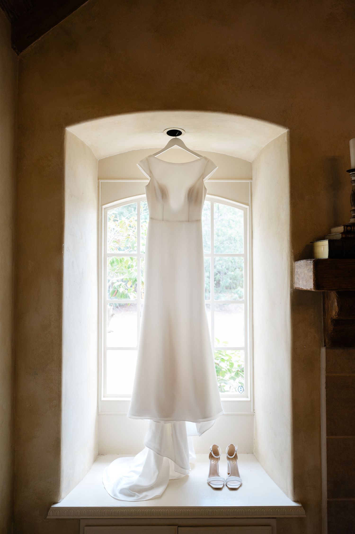 A white wedding dress hangs in an arched window alcove with a pair of bridal shoes below, creating an elegant display.