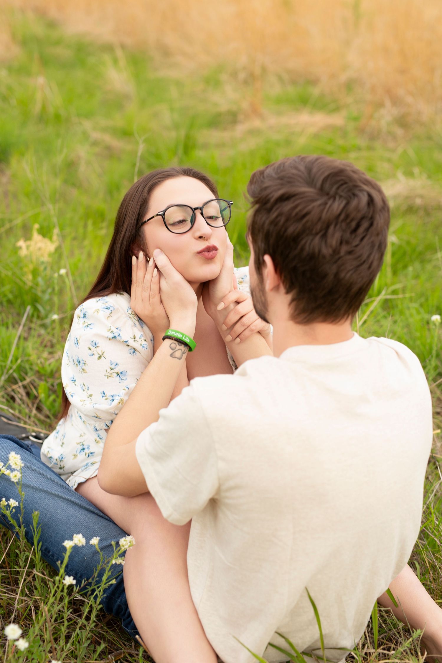Young couple sits together in a grassy meadow on a sunny day, sharing an intimate moment outdoors.