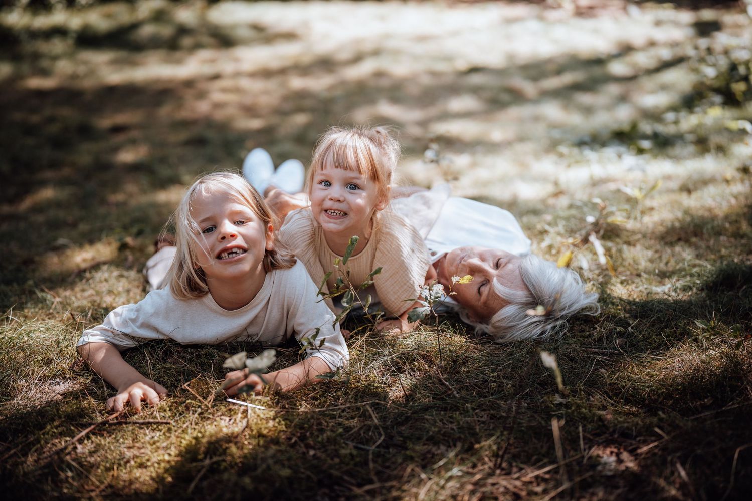 In een zonovergoten bos liggen drie jonge kinderen samen te lachen op de schaduwrijke grond, die bedekt is met gevallen bladeren.