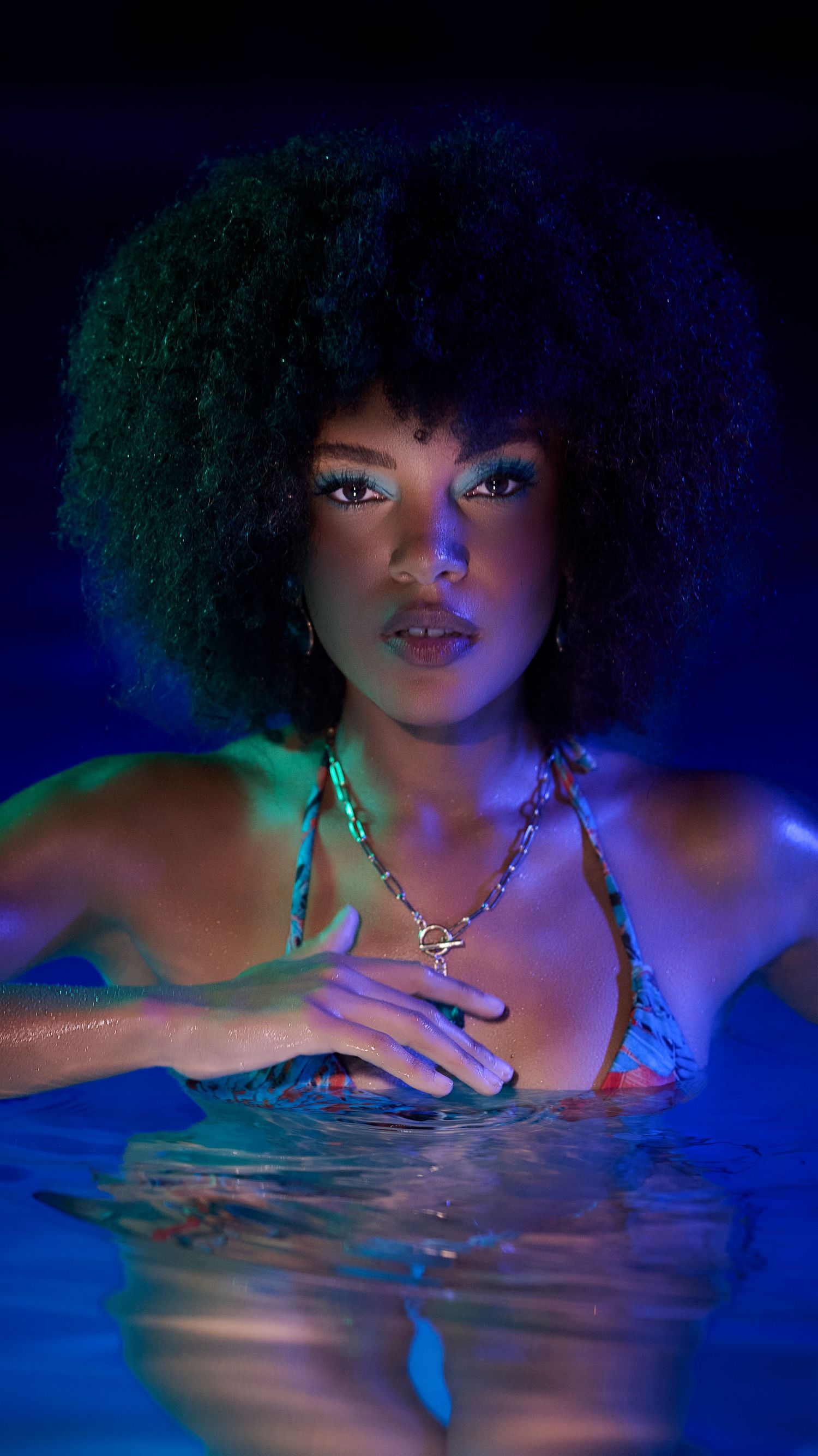 Artistic portrait of a female model in blue and green lighting with dramatic shadows, reflective water, and shimmering jewelry