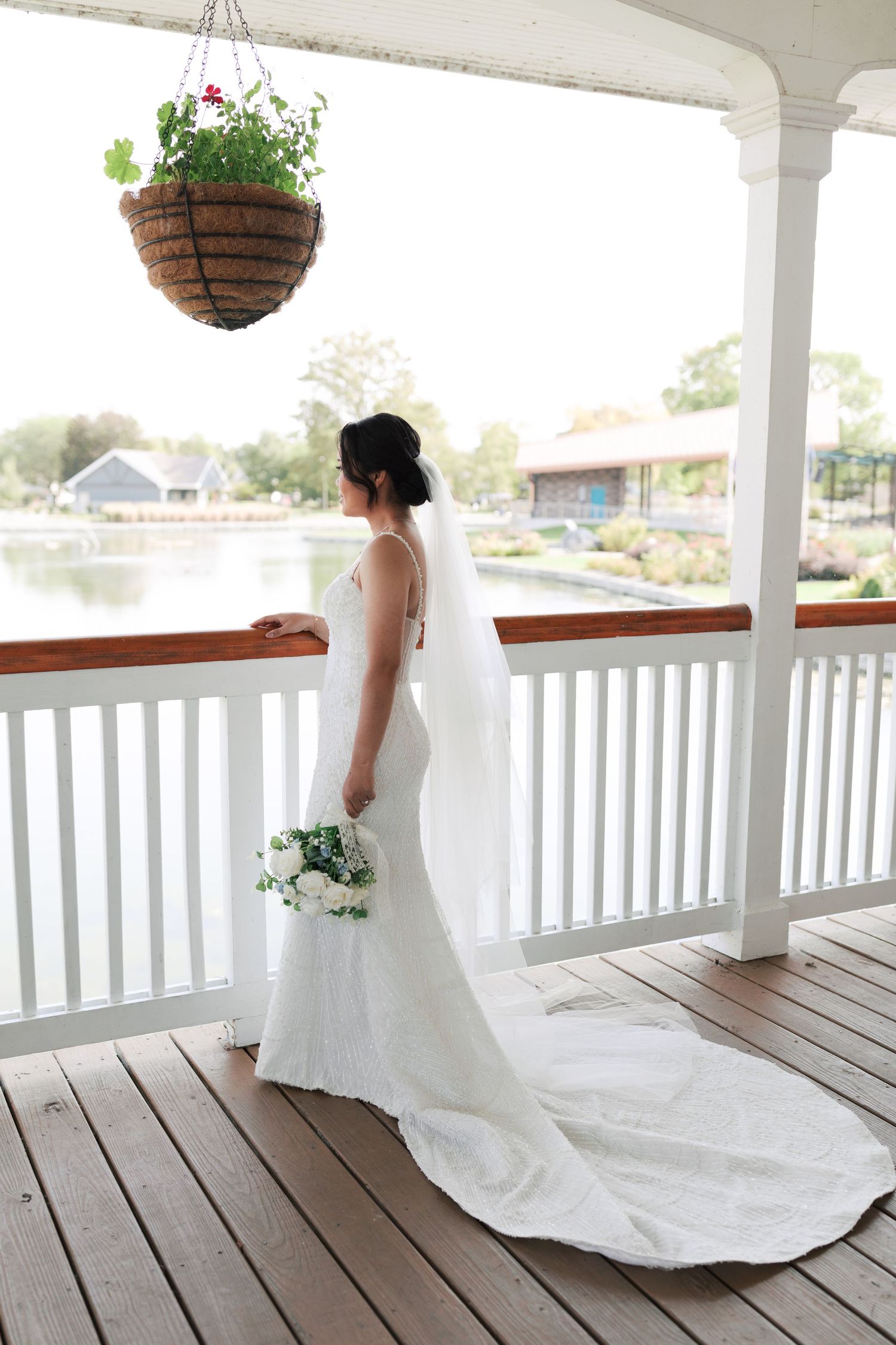 Bride in flowing white gown stands on covered deck overlooking waterfront.