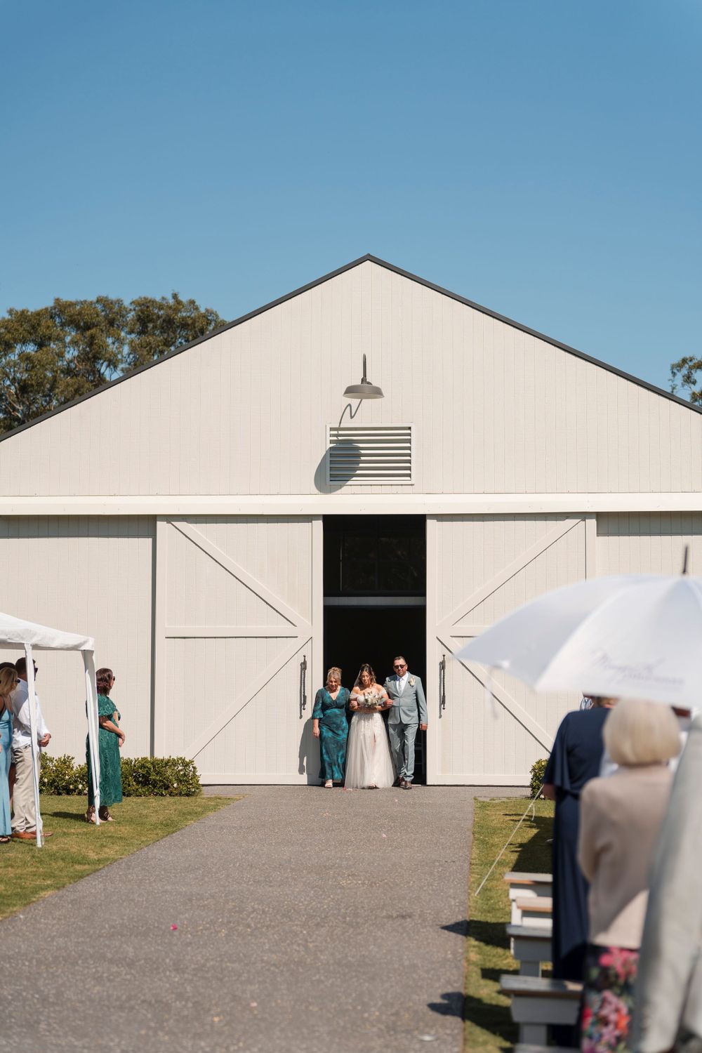 People gather outside a pristine white barn building for a wedding ceremony under clear blue skies.