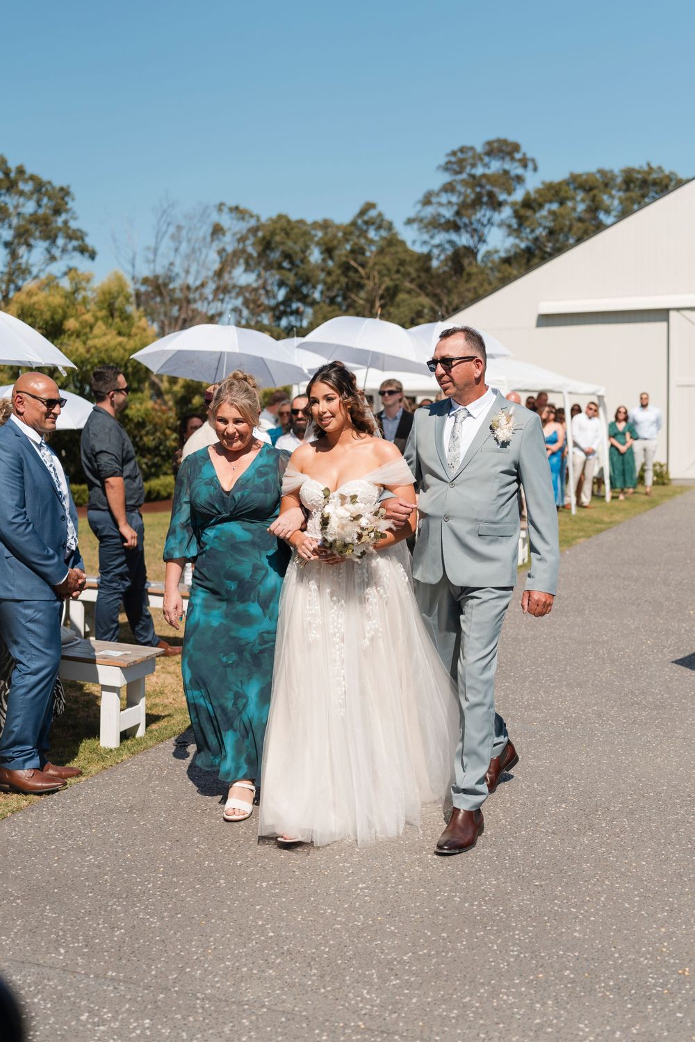 A bride walks down an outdoor path accompanied by guests on a sunny day with white umbrellas visible in the background.